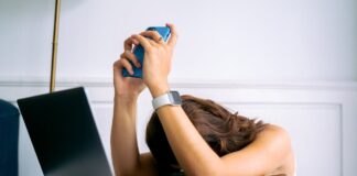 Person sitting at a table with a laptop, holding a phone and showing signs of stress