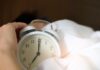 A hand reaching for an alarm clock in a bed