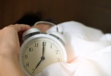 A hand reaching for an alarm clock in a bed