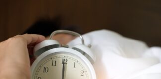 A hand reaching for an alarm clock in a bed