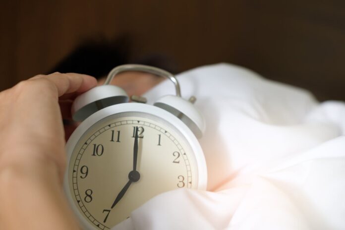 A hand reaching for an alarm clock in a bed