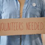 How Outdated Tax Rules Are Quietly Discouraging Volunteer Work Person holding a cardboard sign that says 'VOLUNTEERS NEEDED'