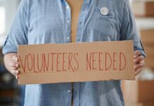Person holding a cardboard sign that says 'VOLUNTEERS NEEDED'