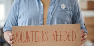 Person holding a cardboard sign that says 'VOLUNTEERS NEEDED'