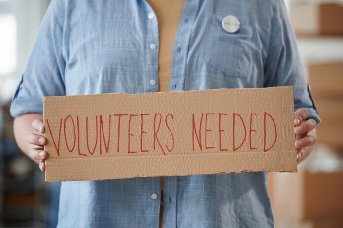 Person holding a cardboard sign that says 'VOLUNTEERS NEEDED'