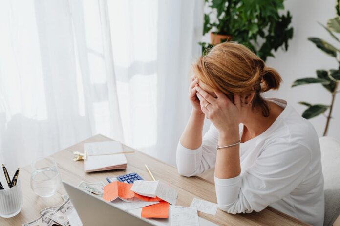 A woman sitting at a desk with her hands on her face, surrounded by paperwork and money, looking stressed