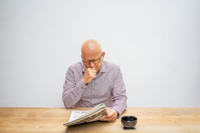 An older man reading a newspaper while sitting at a wooden table with a coffee cup