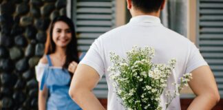 A man hiding a bouquet of flowers behind his back while smiling at a woman
