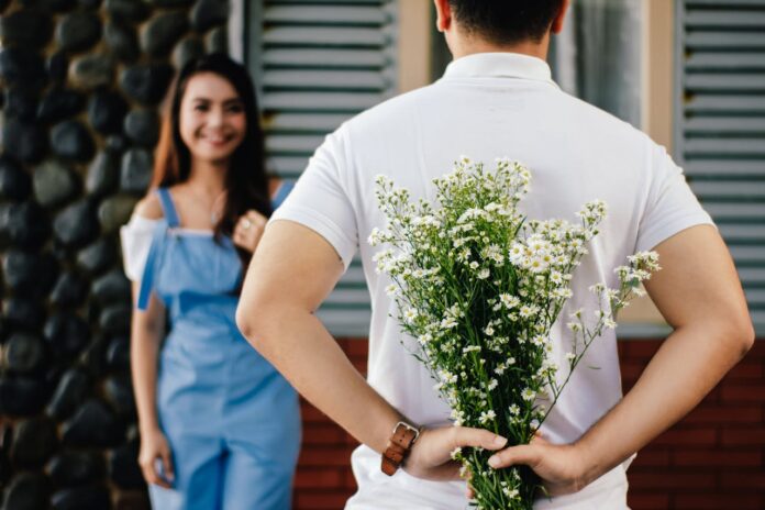 A man hiding a bouquet of flowers behind his back while smiling at a woman